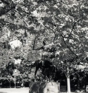 Joan of Arc equestrian statue on a stone pedestal, framed by tree branches and foliage.