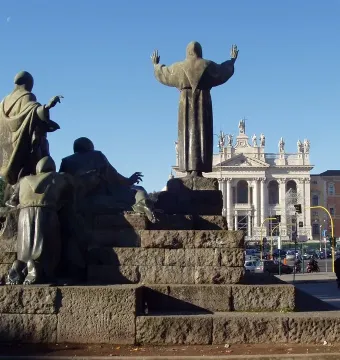 Bronze Monument to Saint Francis of Assisi on a tiered stone pedestal with a church building in the background.