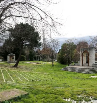 Stone amphitheater seating on a grassy slope with the U Nativu monument and arched gateway in the background.
