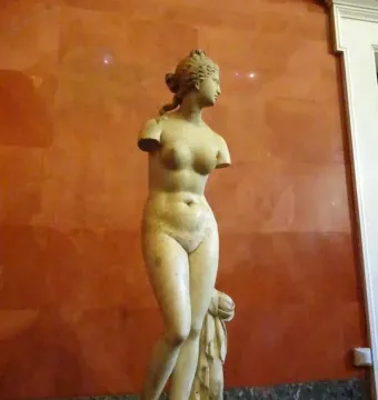 Venus Tauride, a white marble nude female statue with missing arms, standing on a round base against a reddish-orange wall.
