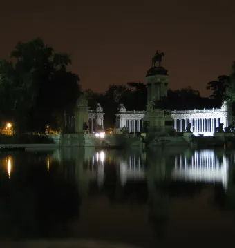 The Alfonso XII Monument lit up at night beside a calm lake, with its colonnade and equestrian statue reflected in the water.