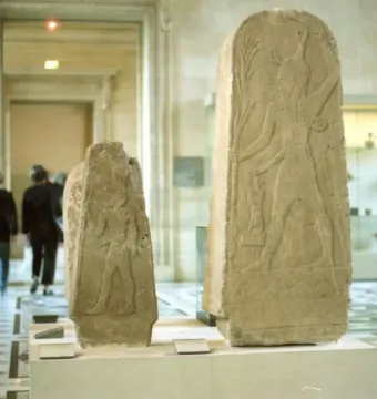 Two carved stelae on display in a museum hall, with visitors walking past in the background.