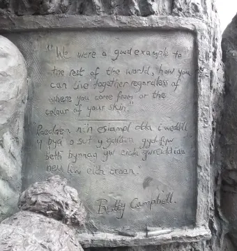 Close-up of the plaque on the Betty Campbell statue with engraved English and Welsh text, framed by sculpted children and a carved wooden trunk.