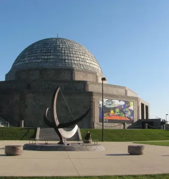 A crescent-shaped sundial sculpture with a tall gnomon stands on a paved plaza in front of a domed building.