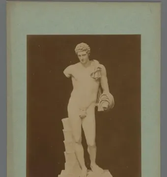 Hermes Belvedere statue standing nude with a cloak over one shoulder, shown on a stepped pedestal in a sepia-toned photograph.
