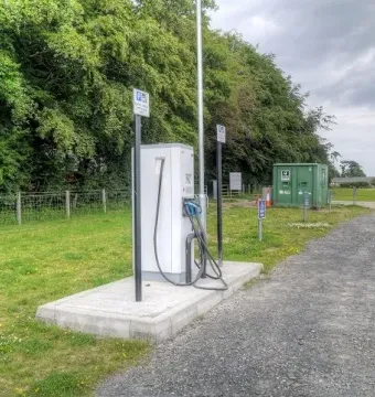 Electric vehicle charging station beside a gravel path at Northumberlandia, with trees, grass, and a utility box behind it.