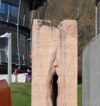 Pi-Chacán, a tall reddish-pink stone sculpture with a deep vertical cleft, standing outdoors near modern buildings and grass.