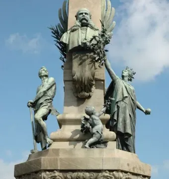 Upper section of the Rius i Taulet Monument with a bronze bust on an obelisk, flanked by bronze figures and a small putto holding a branch.