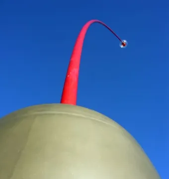 Wind Wand sculpture with a crimson rod rising from a rounded dome against a blue sky.