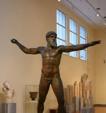 Artemision Bronze, a dark bronze statue of a nude bearded man with outstretched arms in a museum gallery.