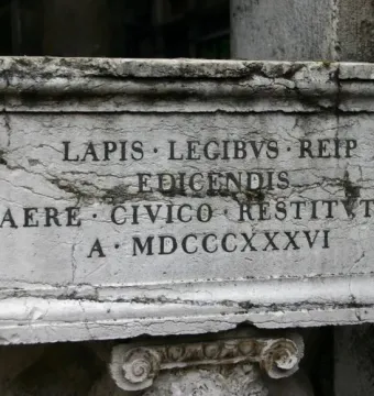 Weathered stone plaque with a carved Latin inscription and a decorative stone capital beneath it.