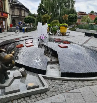 Reksio Monument beside a split egg-shaped fountain with pink-lit water in a town square.