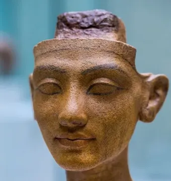 Unfinished head of Nefertiti in reddish-brown stone with a long neck and carved facial features on a light speckled base.