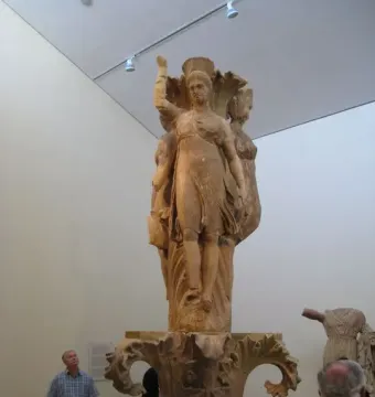 Dancers of Delphi sculpture with three draped female figures on an acanthus column in a museum gallery.