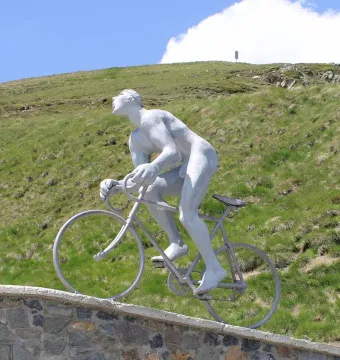 Le Géant du Tourmalet sculpture of a nude male cyclist climbing on a stone wall with mountains and blue sky behind it
