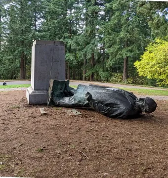 Toppled Harvey W. Scott statue lying on its side beside a stone pedestal in a park with tall pine trees.