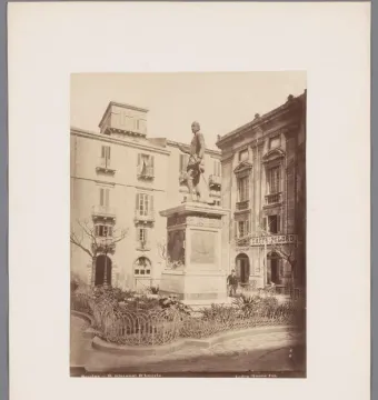 The John of Austria monument stands on an ornate pedestal in a public square, surrounded by a low decorative fence and garden.