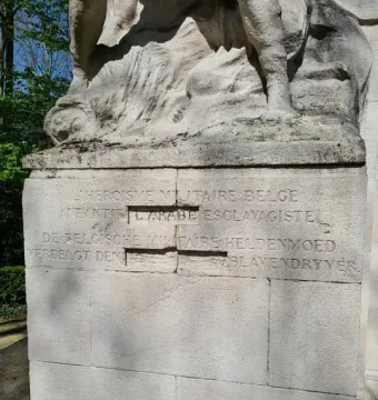 Close-up of the Monument to the Belgian Pioneers in Congo showing carved French and Dutch inscriptions on stone blocks beneath a weathered sculpture fragment.