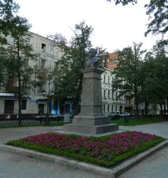 Bust of Pushkin atop a tall stone monument beside a flowerbed of pink blooms, with city buildings and trees behind it.