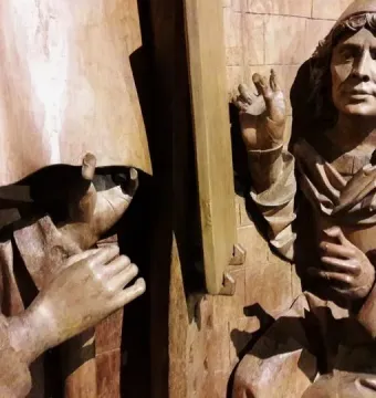 Saint Luke wooden sculpture detail showing two draped figures gesturing beside a paneled wooden background.