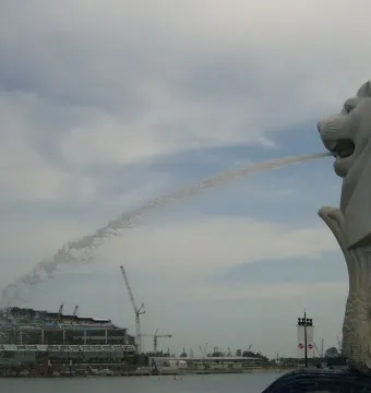 Merlion statue spraying water into the bay with a modern building and cranes in the background.