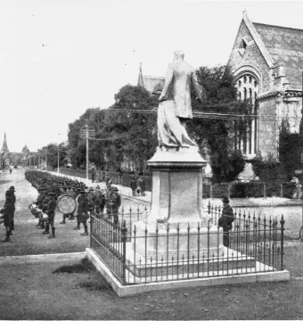 Rolleston Statue on a tall pedestal beside a fenced street, with uniformed men marching in the distance.