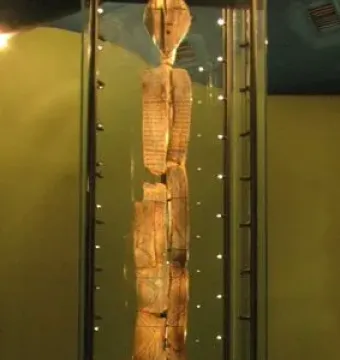 The Shigir Idol stands upright in a glass case, showing an oval head and carved geometric patterns on its weathered wooden surface.