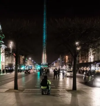 The Spire of Dublin illuminated in blue-green light at night on a city street.