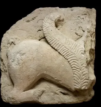 Weathered stone fragment of the El Salobral Sphinxes showing the hindquarters and curved body of an animal with a textured tail arched over its back.