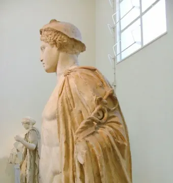 Hermes Criophorus marble statue in profile with a ram beside him, wearing a cap and draped garment.