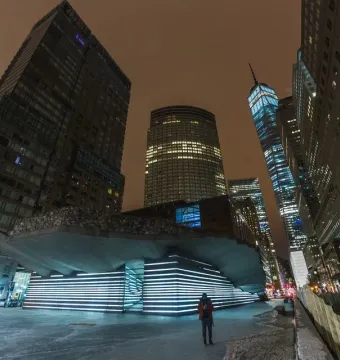 Irish Hunger Memorial with a rocky upper facade and illuminated stepped section beside downtown skyscrapers at night.