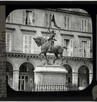 Joan of Arc equestrian statue on a tall pedestal in front of a building with arched shops and shuttered windows.