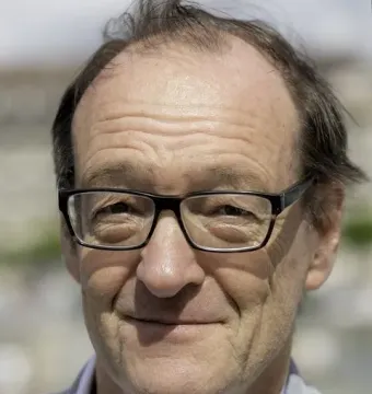 Smiling older man with black-rimmed glasses wearing a light jacket and white collared shirt.