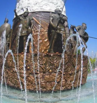 Bronze base of the Warrior on Horseback monument with armored warriors, spears, shields, and surrounding fountain jets.