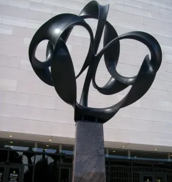 Continuum, a tall abstract metal sculpture with looping ribbons on a granite pedestal outside a modern building.