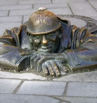 Čumil, a bronze man wearing a helmet, leans out of a manhole in a paved street.