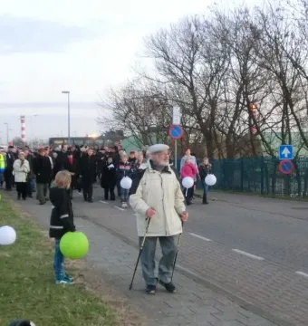 A procession of people walks along a paved path near the Kindertransport monument, carrying glowing white and green lanterns.