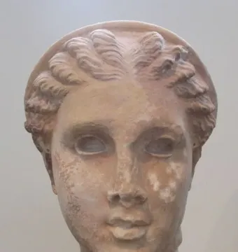 Weathered stone head of Lycosoura Artemis on a pedestal, with braided hair and a wreath.
