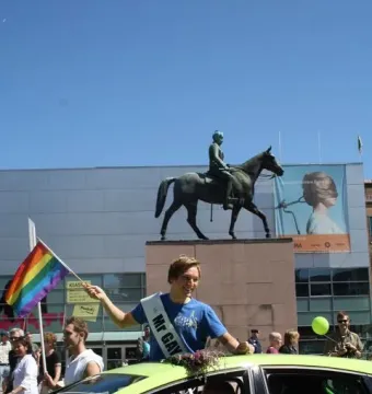 Marshal Mannerheim equestrian statue on a granite plinth with a crowd and a person in a lime green car holding a rainbow flag in the foreground.