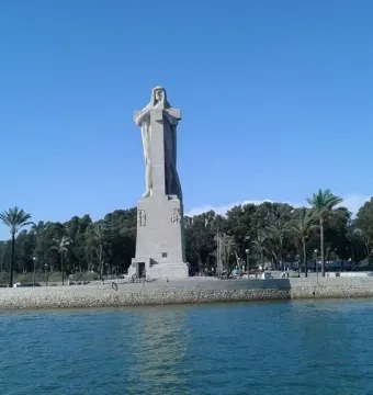 Monument to Columbus rising from a stone-walled platform surrounded by water under a bright blue sky.