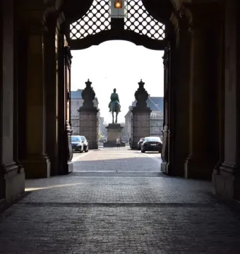 Christian IX equestrian statue seen through a dark stone archway, with a sunlit plaza and parked cars beyond.