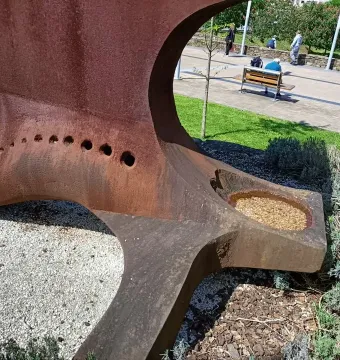 Dorna sculpture with a rusty metal surface, circular holes, and a basin filled with gravel in a park setting.
