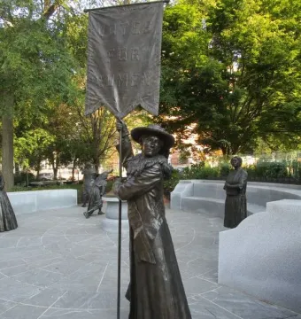 Virginia Women’s Monument sculpture with a central figure holding a tall Votes for Women banner, with additional statues on a stone patio behind it.