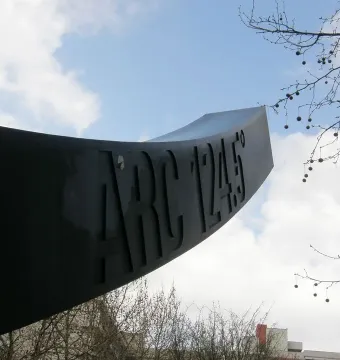 Arc de 124,5° sculpture seen from below against a partly cloudy blue sky, with bare branches framing the upper right.