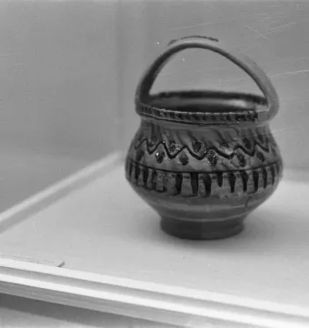 A small ornate ceramic vessel with a handle and wavy decorative patterns, displayed in a clear case.