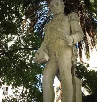 The Count of Porto Alegre monument, a light-colored military figure sculpture with epaulets and a sash, seen from below among trees and palm fronds.