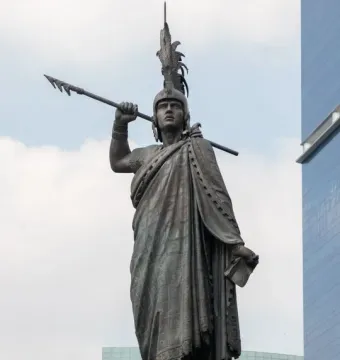 Cuauhtémoc Monument statue with a feathered headdress, spear, and document against a cloudy sky.