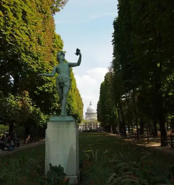 The Greek Actor statue on a tall stone pedestal in a tree-lined park, with a grassy avenue and distant domed building behind it.