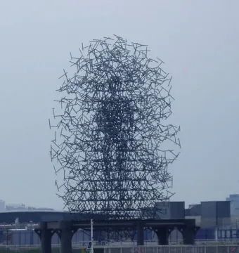 Quantum Cloud sculpture of thin dark metal rods forming a large oval shape beside the water.