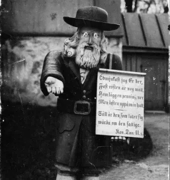 Rosenbom, a wooden bearded figure in a hat holding out one hand and a sign.